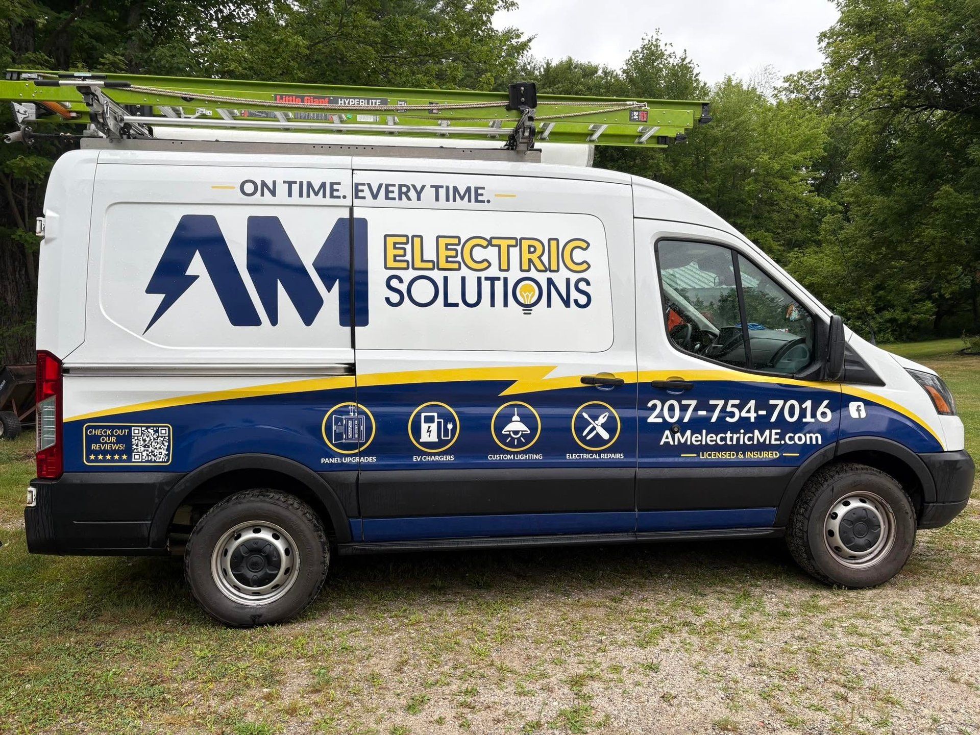 White and blue AM Electric Solutions service van parked on grass with company logo, tagline, phone number, and service icons displayed on the side.